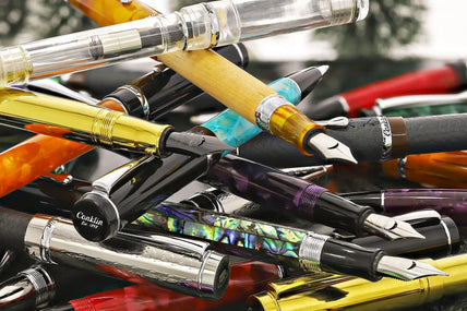Multi-colored fountain pen collection with translucent and metallic accents, featuring a variety of vibrant hues including rainbow-like iridescence.